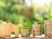 Stapels munten met plantjes die eruit lijken te groeien met daarnaast een stuk hout in de vorm van een huis